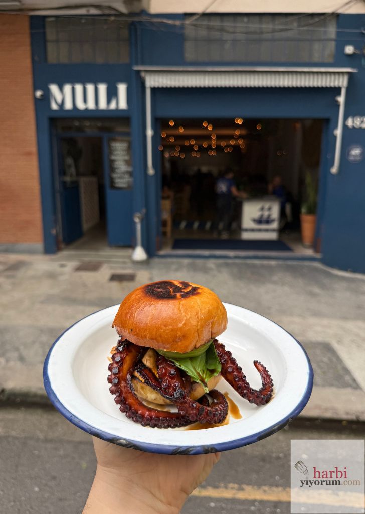 Muli’nin mavi cephesi önünde, ekmeğin kenarlarından ahtapot kolları taşan burger