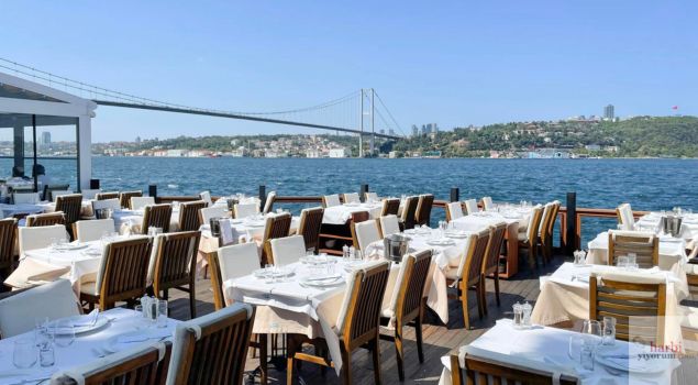 Boğaz Köprüsü manzaralı, deniz kenarında açık hava oturma alanı sunan şık bir restoran. Beyaz masa örtüleriyle düzenlenmiş masalar, İstanbul Boğazı'nın büyüleyici atmosferi eşliğinde romantik ve özel bir yemek deneyimi için ideal bir ortam sunuyor.