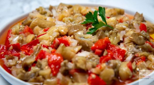 Bir tabakta sunulan közlenmiş patlıcan salatası. Közlenmiş patlıcanlar, kırmızı biber parçaları ve ince doğranmış soğanlarla karıştırılmış. Üzerinde taze maydanoz yaprağı ile süslenmiş, parlak zeytinyağı sosuyla lezzetlendirilmiş. Canlı renkler ve taze malzemelerle iştah açıcı bir görünüm sunuyor.