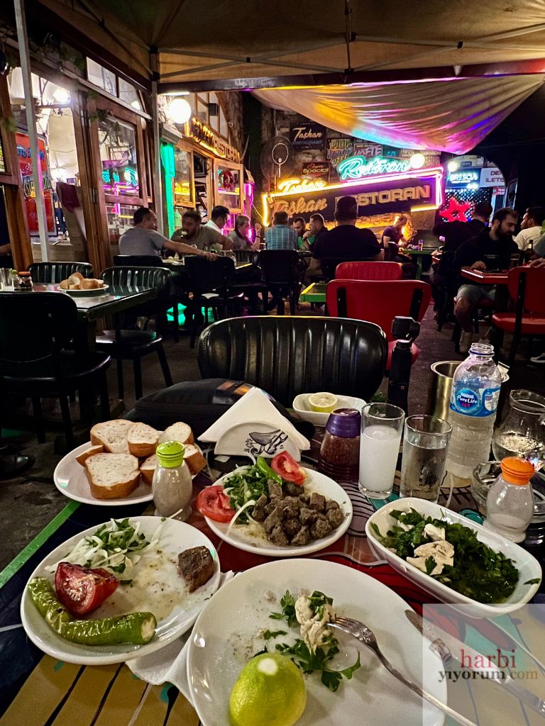 Taşhan Restoran Bartın'da meyhane masası, üzerinde meze tabakları, rakı bardakları ve ekmek dilimleri. Arka planda otantik dekorasyon ve neon ışıklarla süslenmiş restoranın dış bölümü.